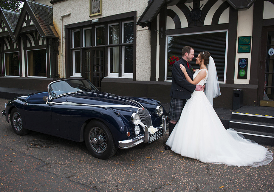 Black wedding car outside hotel with bride and groom embracing in front